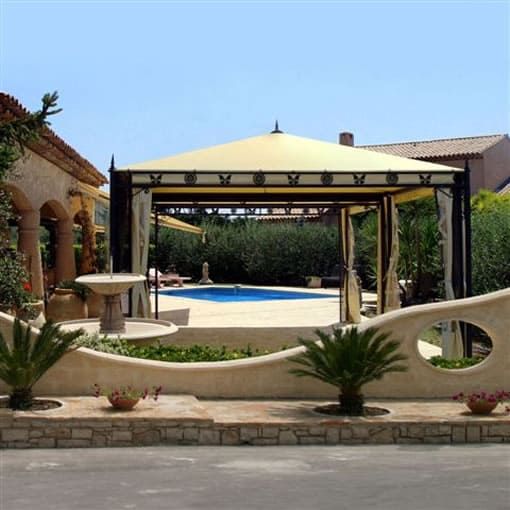 Gazebo con vista sulla piscina, tetto in tela beige, muro in pietra, piante, giornata di sole.