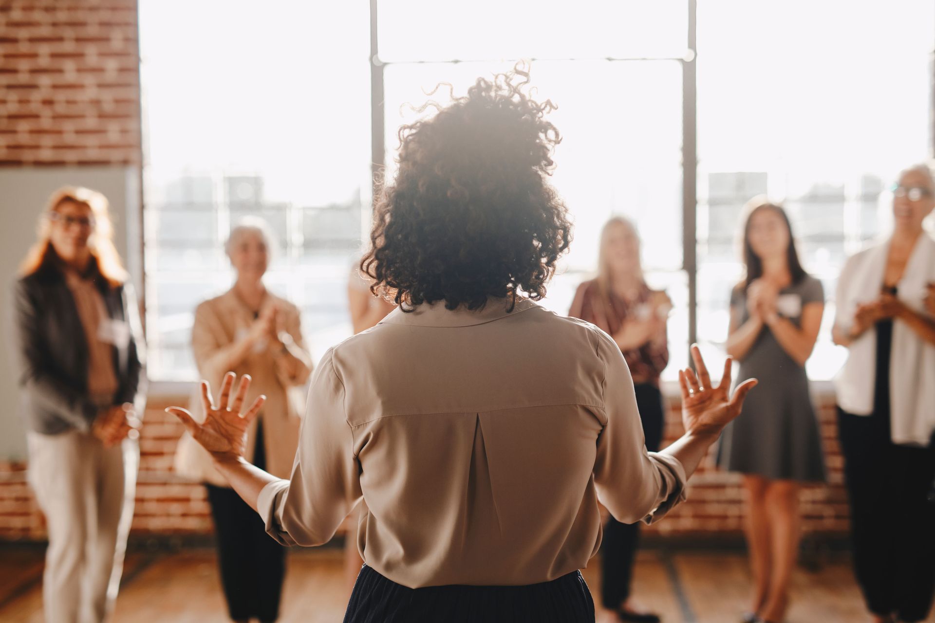 Woman speaking to a group of applauding women in a bright room, back view.