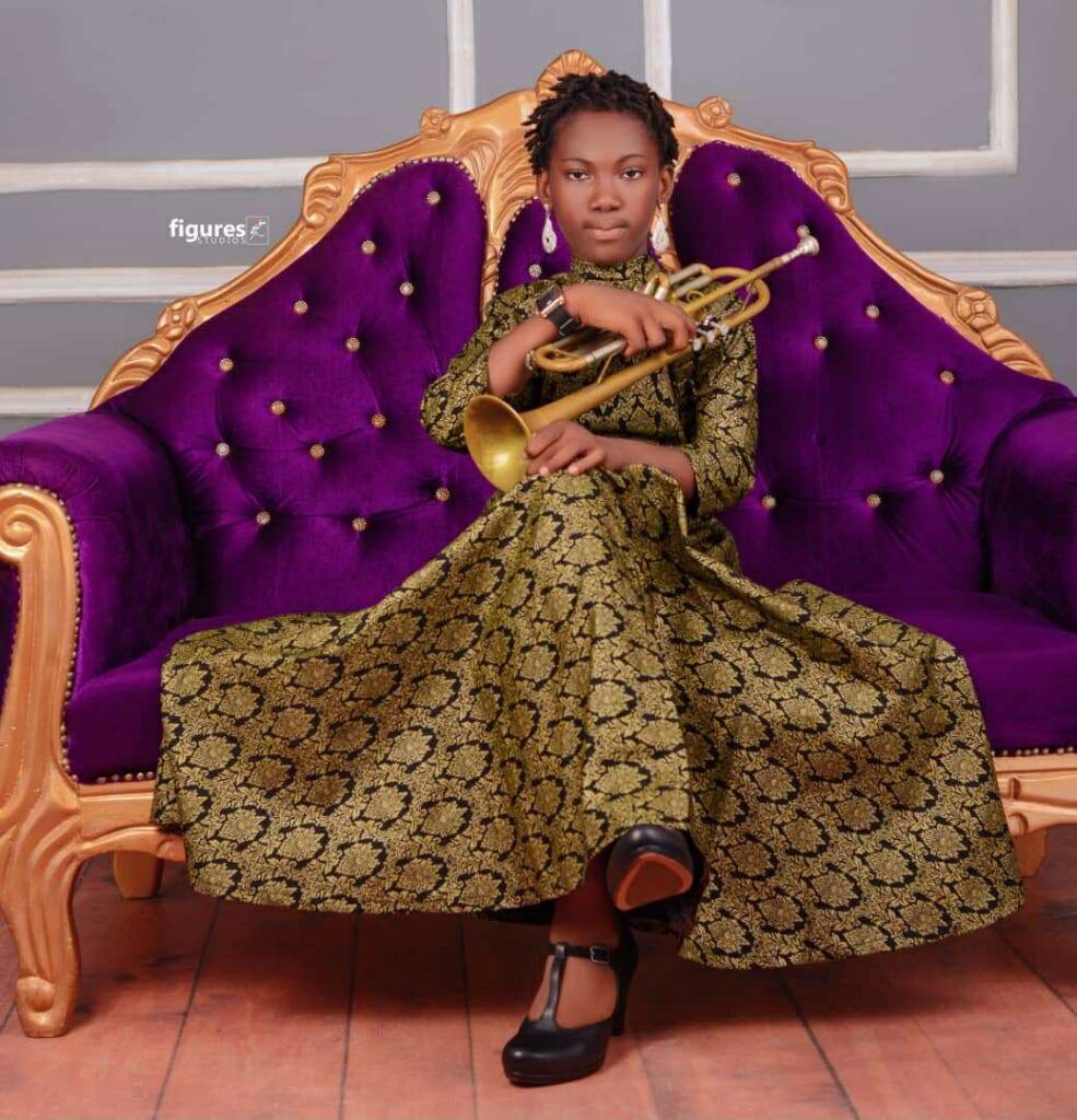Young person in a formal dress holding a trumpet, seated on a purple velvet throne.