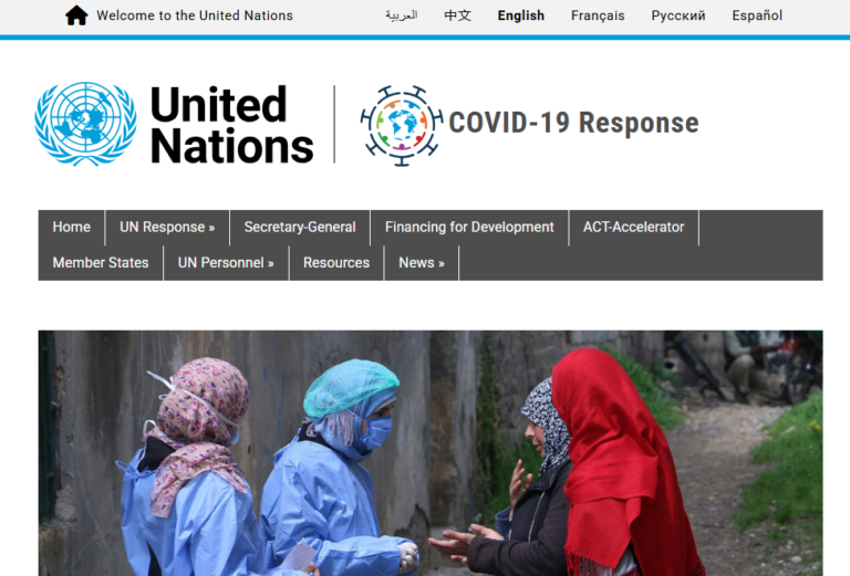 Medical workers in protective gear speak with women outdoors, likely in a refugee camp.