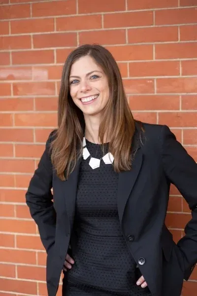 Woman in black blazer and dress smiles, hands on hips, against brick wall.