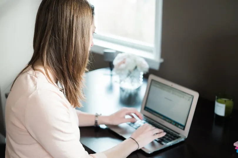 Woman typing on a laptop at a dark desk near a window; a bouquet and candle are nearby.