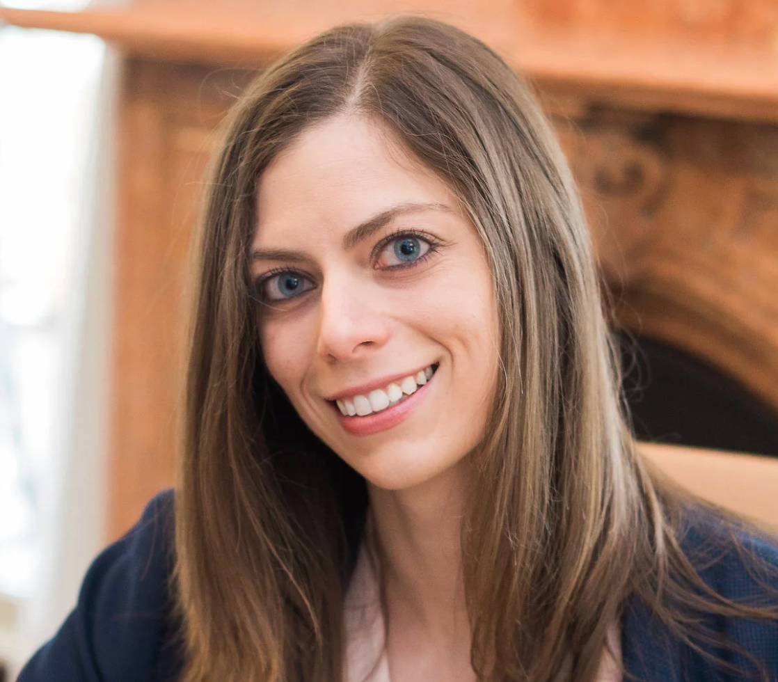 Woman with long brown hair smiles at the camera in front of a fireplace.