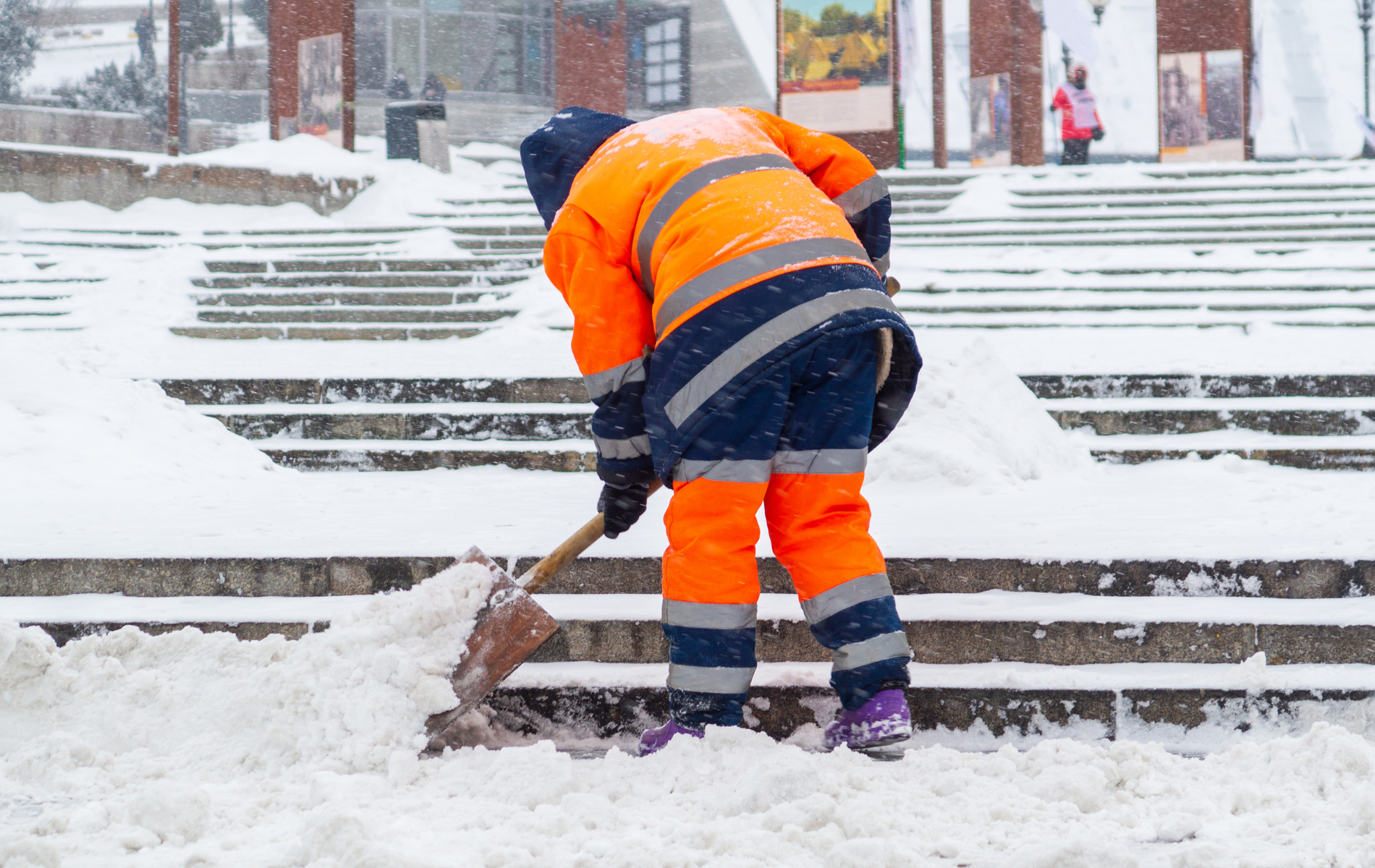 Person in orange workwear shoveling snow off outdoor steps.