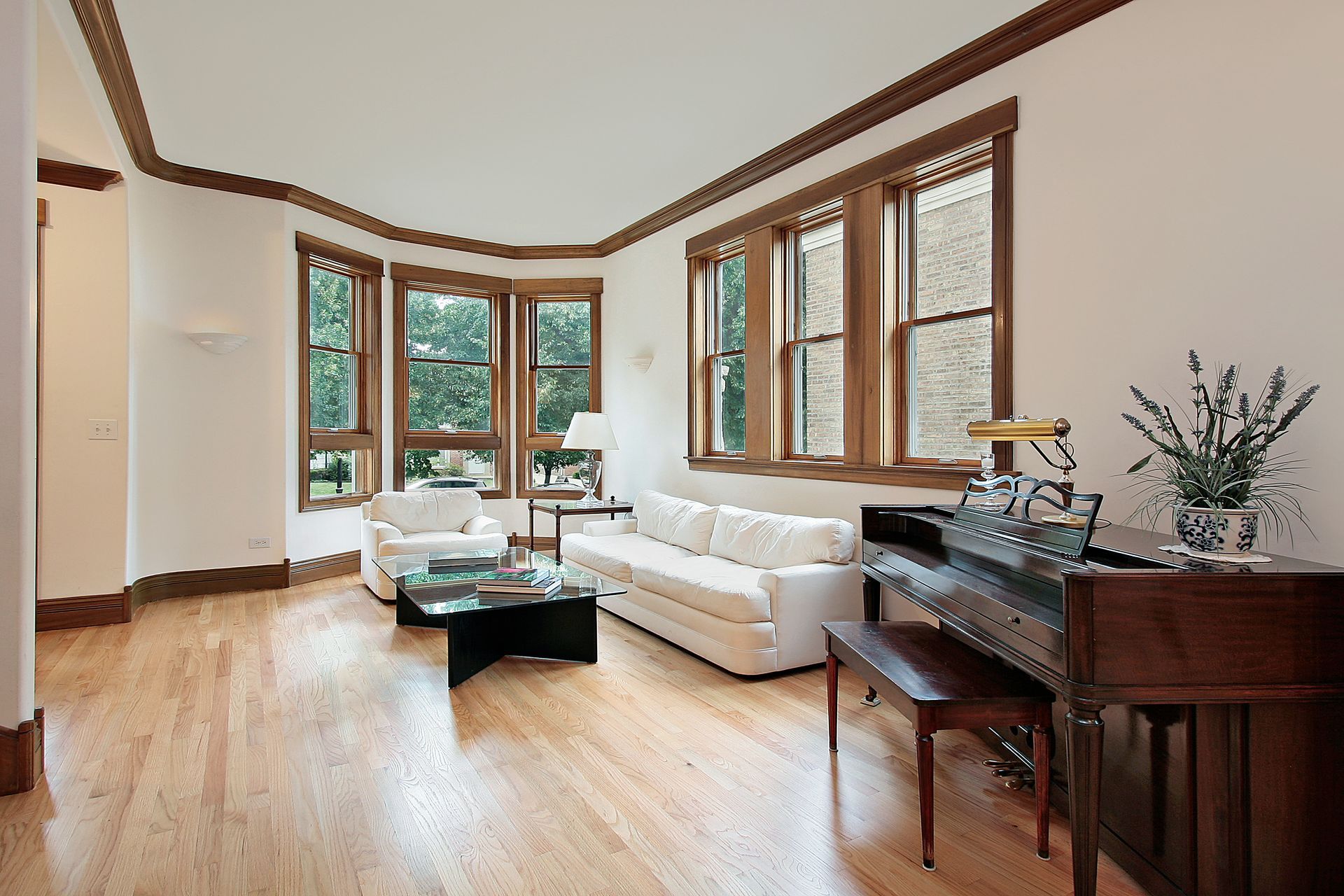 Living room with hardwood floors, white walls, and a piano. Windows with wood trim, and white sofa and chairs. Living room with hardwood floors, white walls, and a piano. Windows with wood trim, and white sofa and chairs.