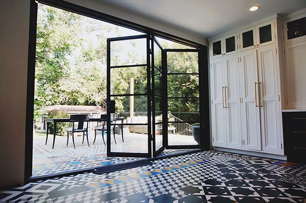 Open black-framed glass doors to patio; patterned tile floor, white cabinets with gold handles. Open black-framed glass doors to patio; patterned tile floor, white cabinets with gold handles.