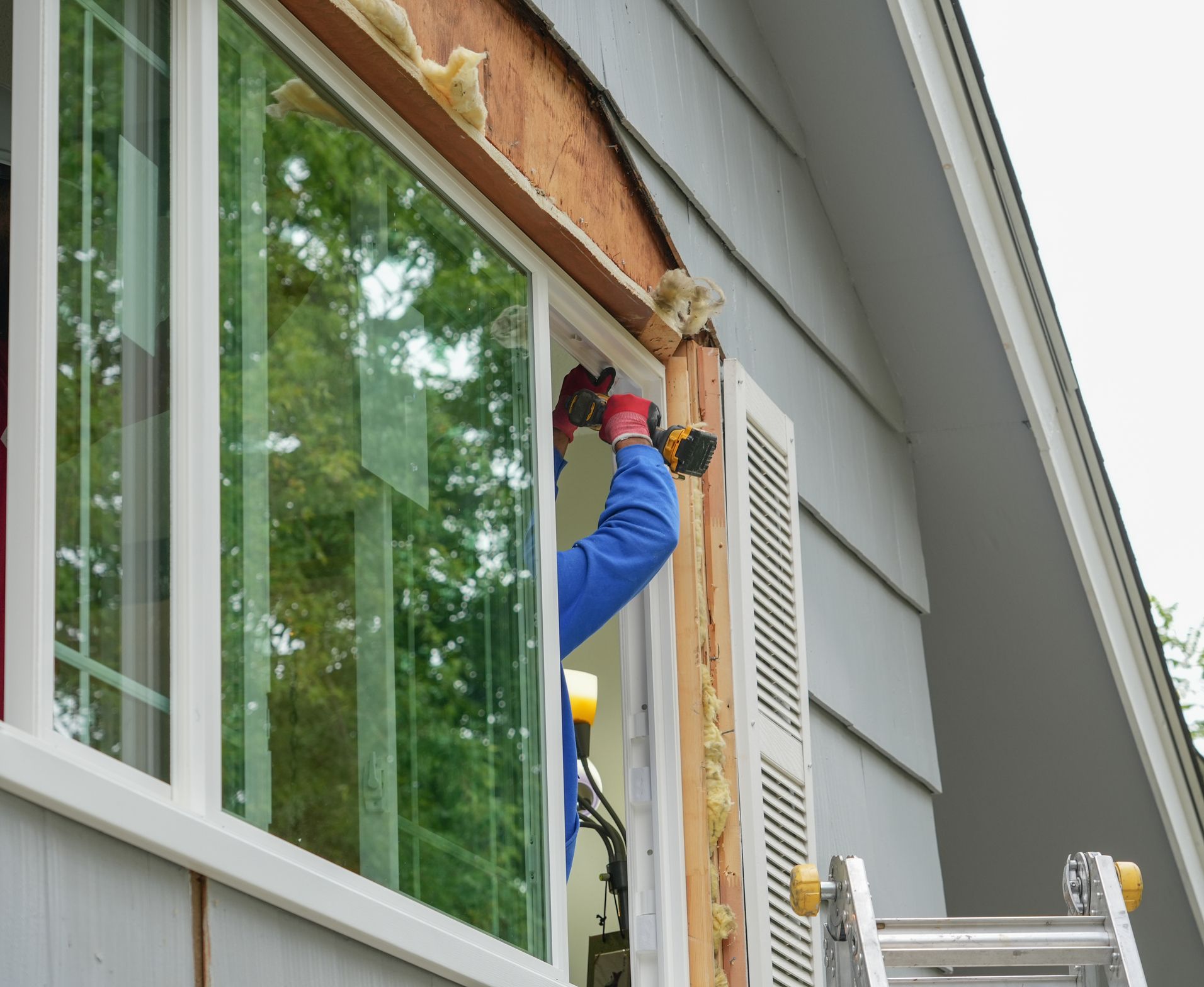 Person installing a window, using a drill on a home's exterior with gray siding and a ladder.