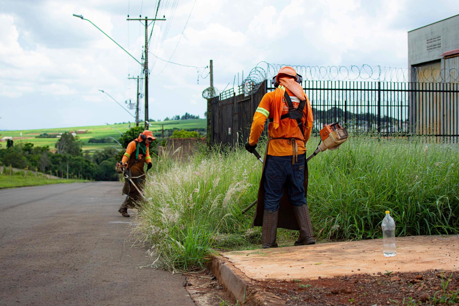 Two workers in orange safety gear use string trimmers to cut tall grass along a paved road near a fenced industrial site.