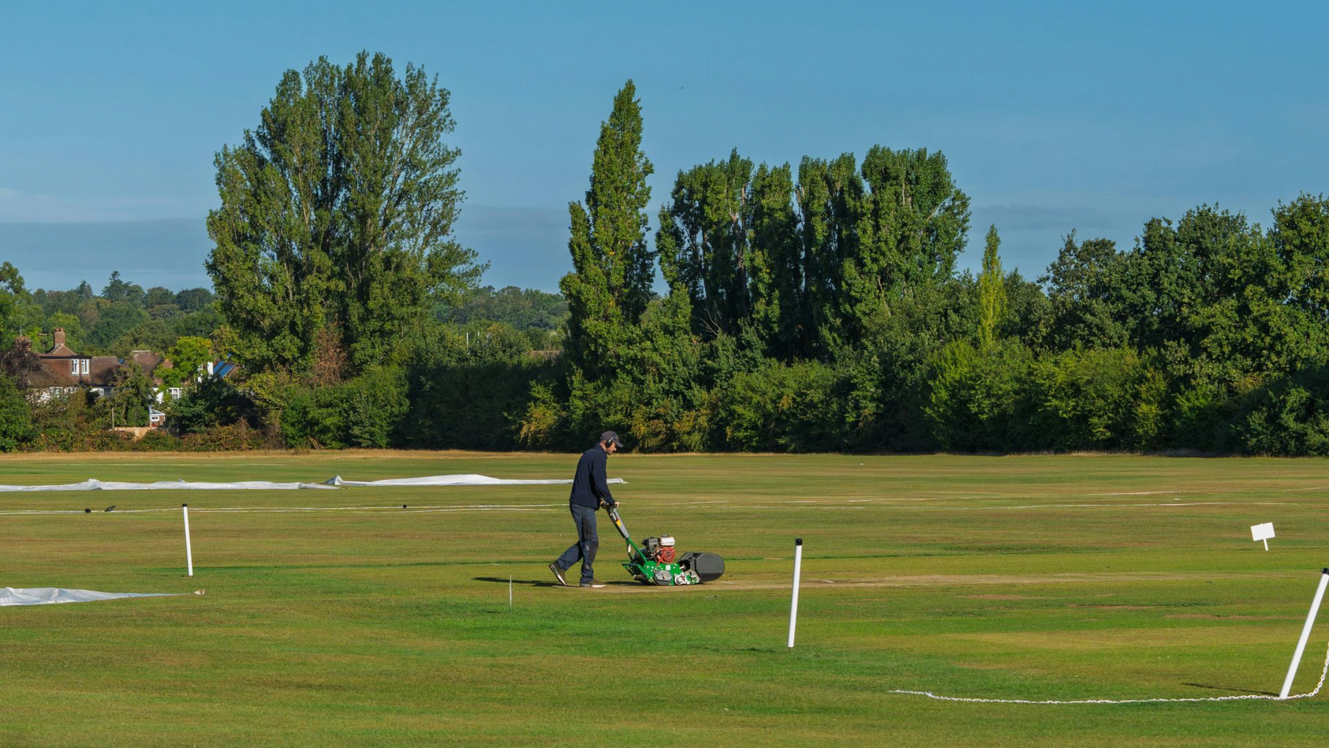 Person mowing a grassy field with white marker posts and trees in the background
