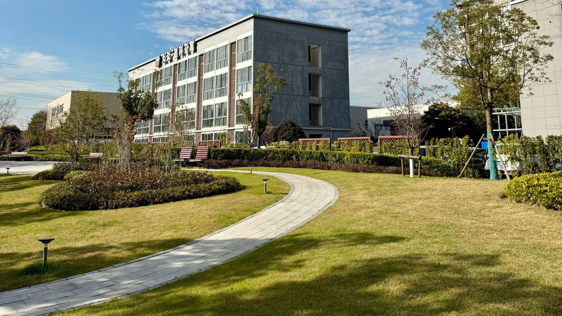 A curving stone path leads through a grassy park toward a multi-story, modern gray building under a sunny blue sky.
