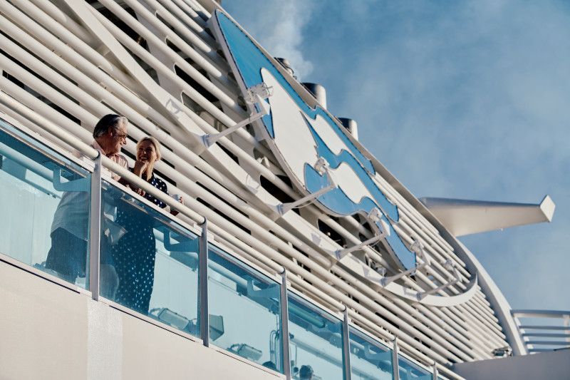 Couple on a cruise ship balcony, blue skies, white ship exterior with a logo.