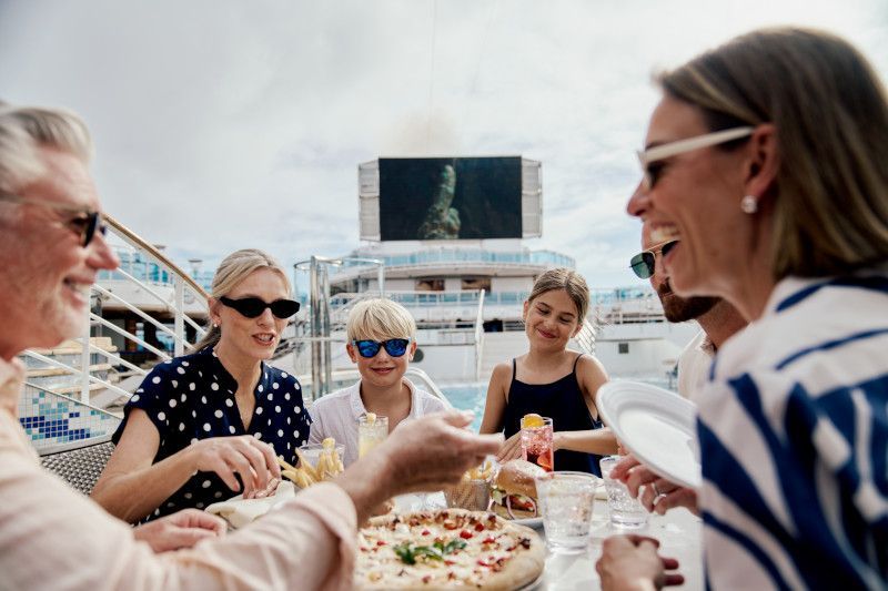 Family laughing while eating pizza on a cruise ship deck, with a large screen in the background.