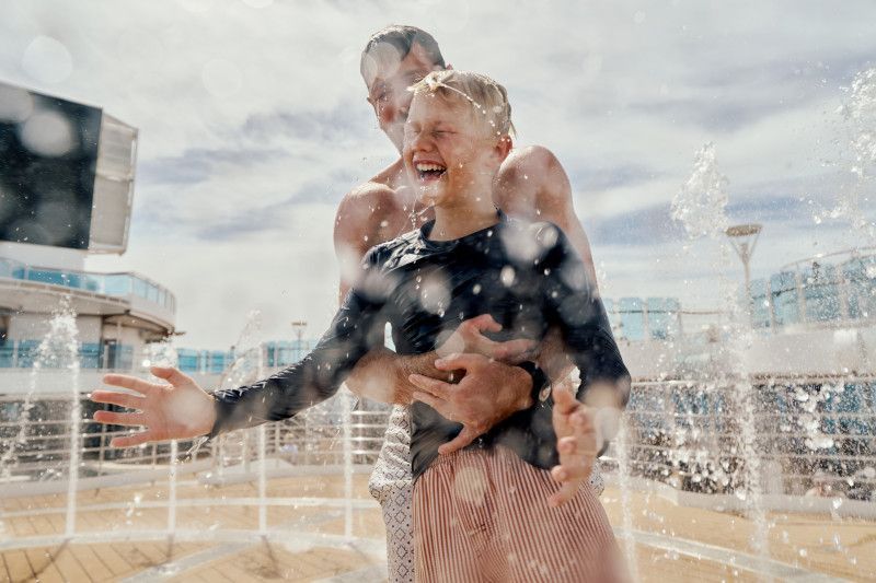 Man splashing woman in water fountains, both laughing on a sunny deck.