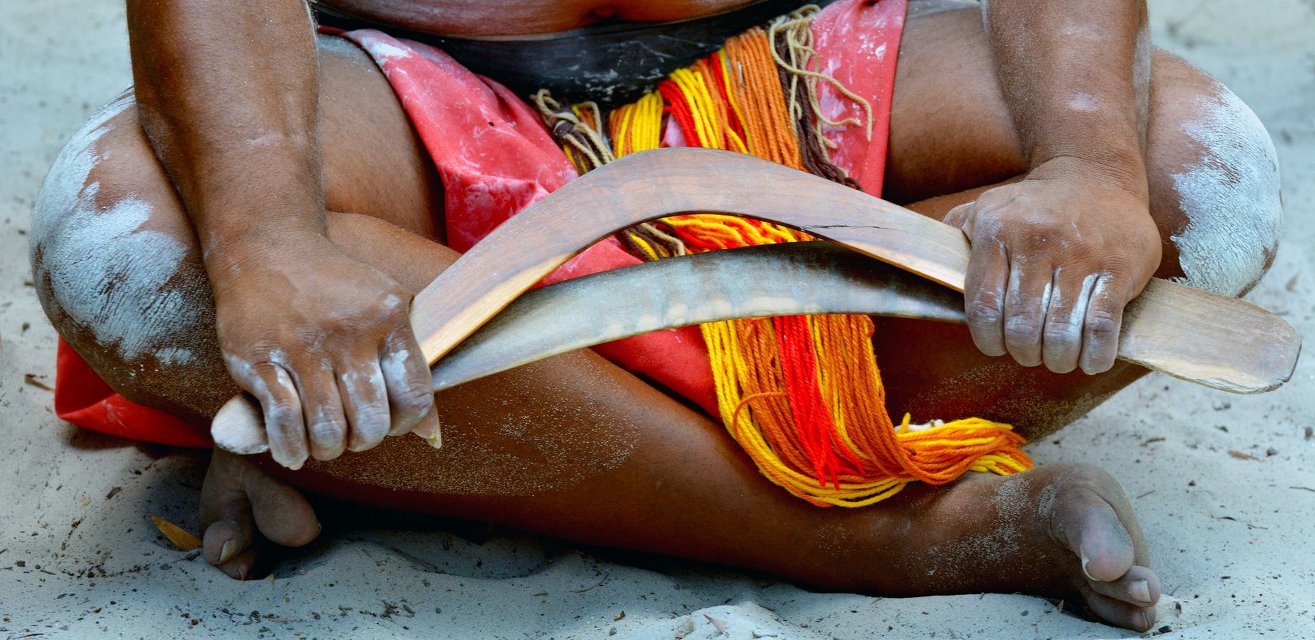 A person holding a boomerang, sitting cross-legged on sand, wearing red and yellow attire.