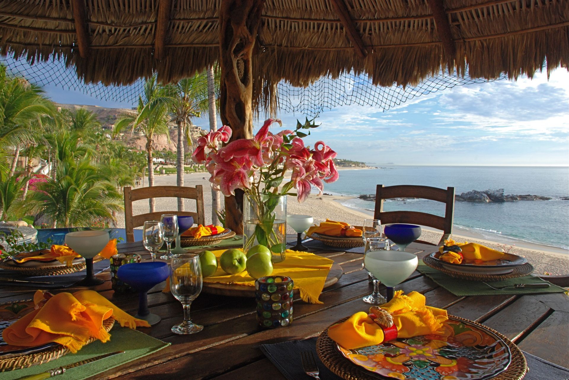 Table set for meal with ocean view, under thatch roof. Flowers, food, and drinks on the table.