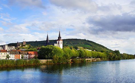 Town along a river, featuring buildings with spires, a green hill, and blue water under a cloudy sky.