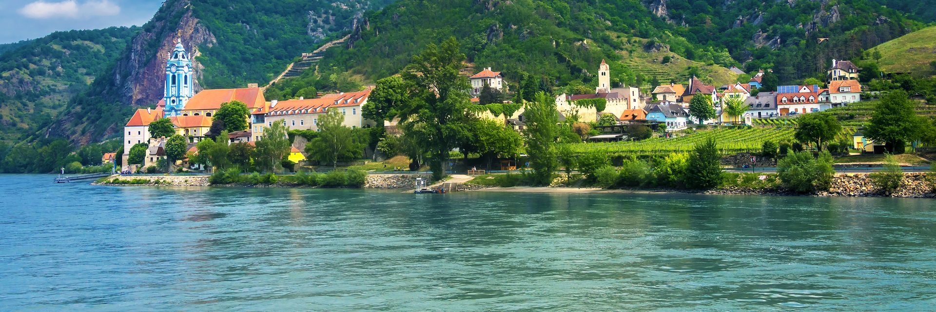A scenic riverside village with colorful buildings, lush trees, and a church against a mountain backdrop.