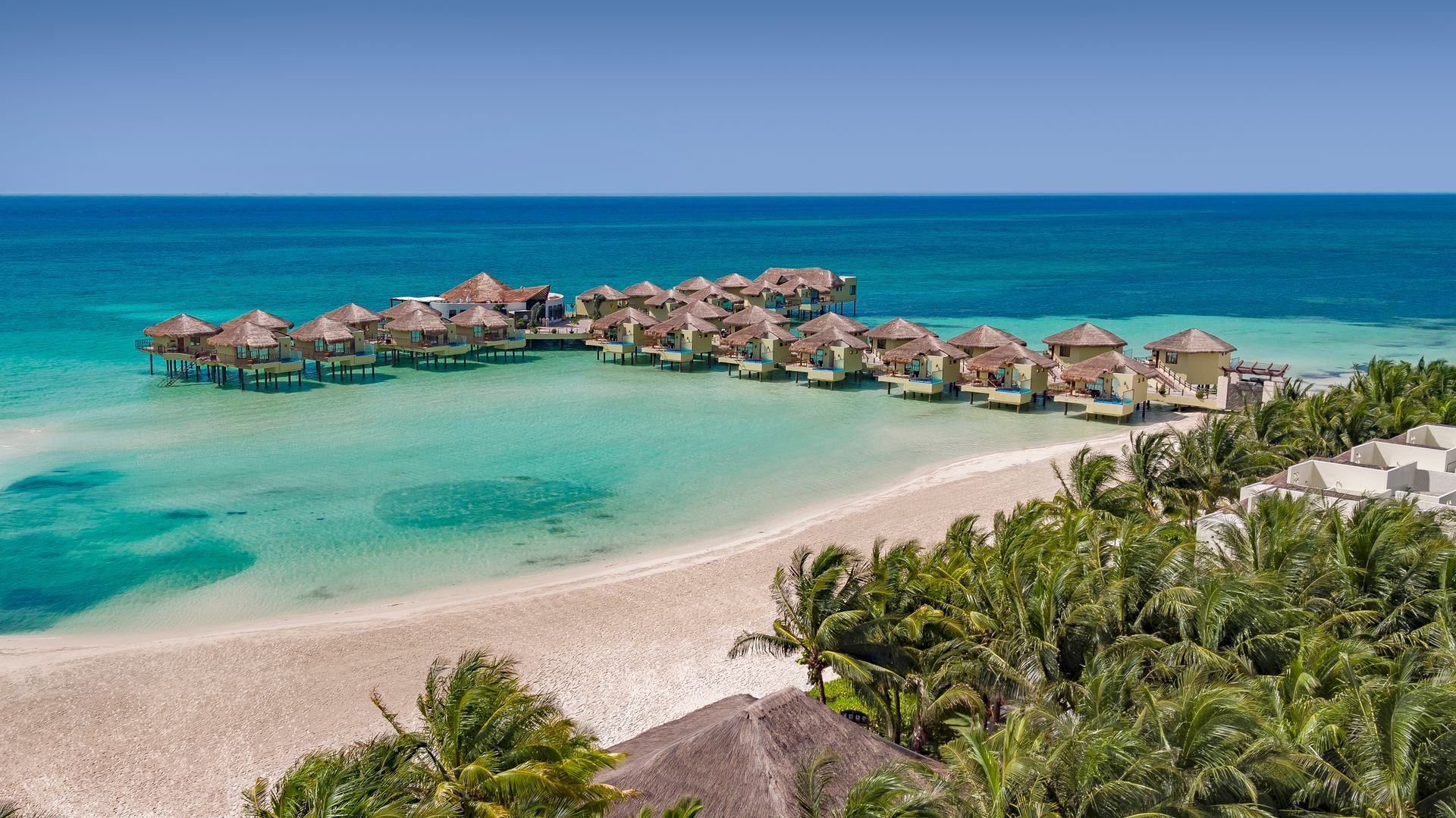 Overwater bungalows on a turquoise lagoon. White sand beach with palm trees under a clear blue sky.