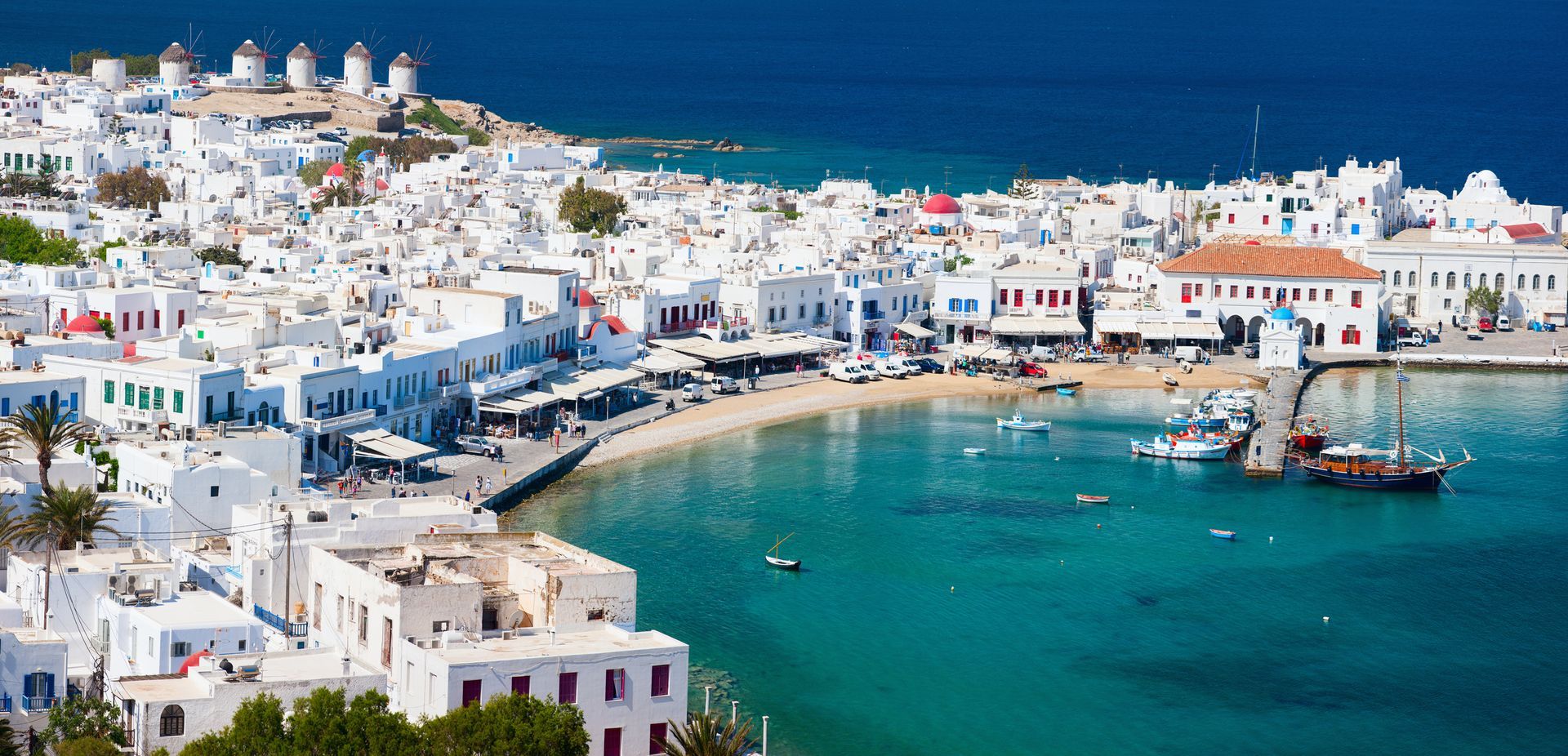 White buildings with blue accents line a seaside town with boats in the turquoise water.