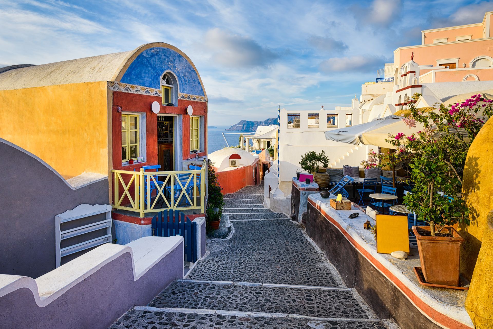 Cobblestone street in Santorini, Greece, with colorful buildings, ocean view, and outdoor cafe.