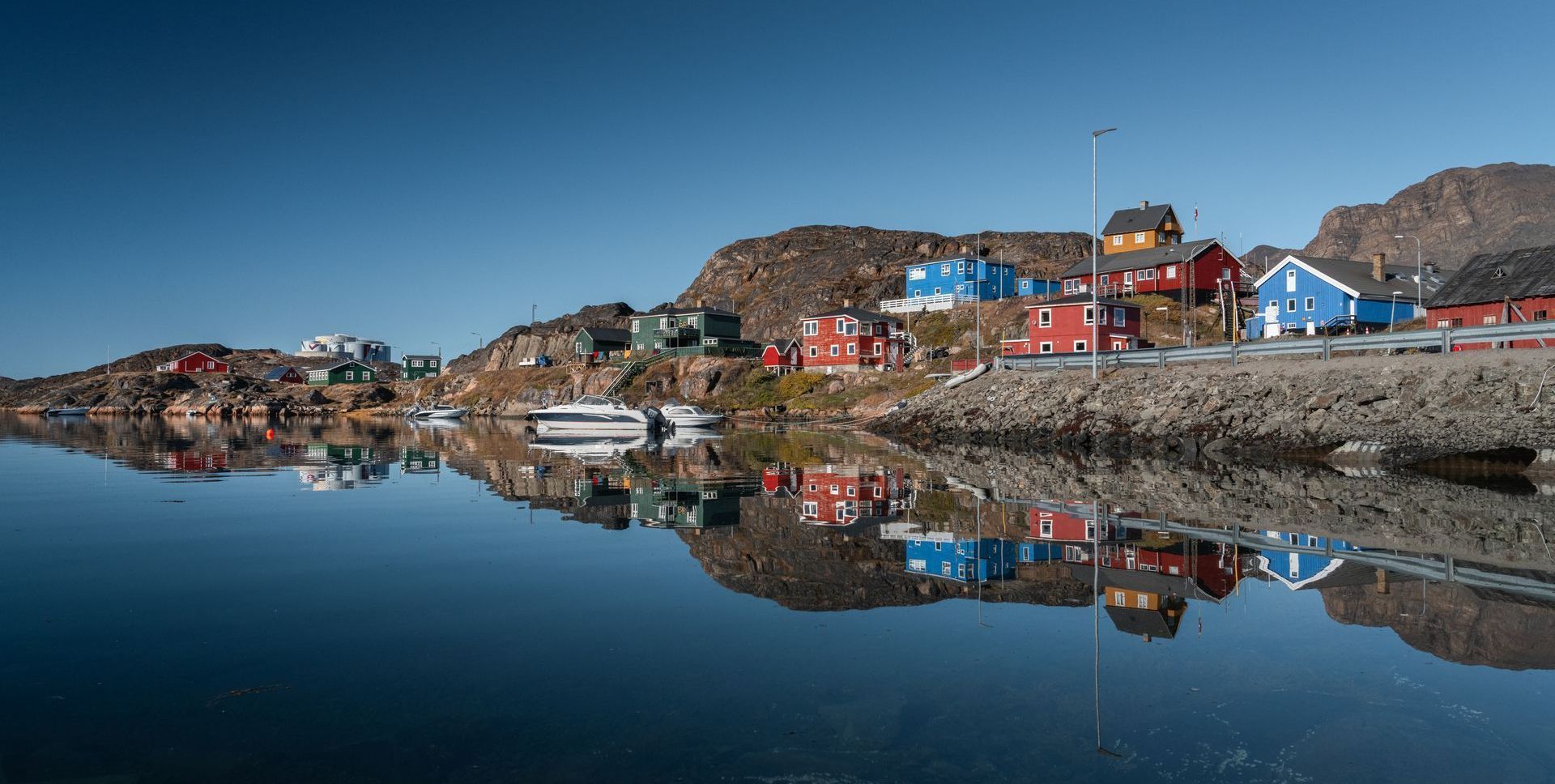 Colorful houses line a rocky shore, reflected in the still, blue water beneath a clear sky.