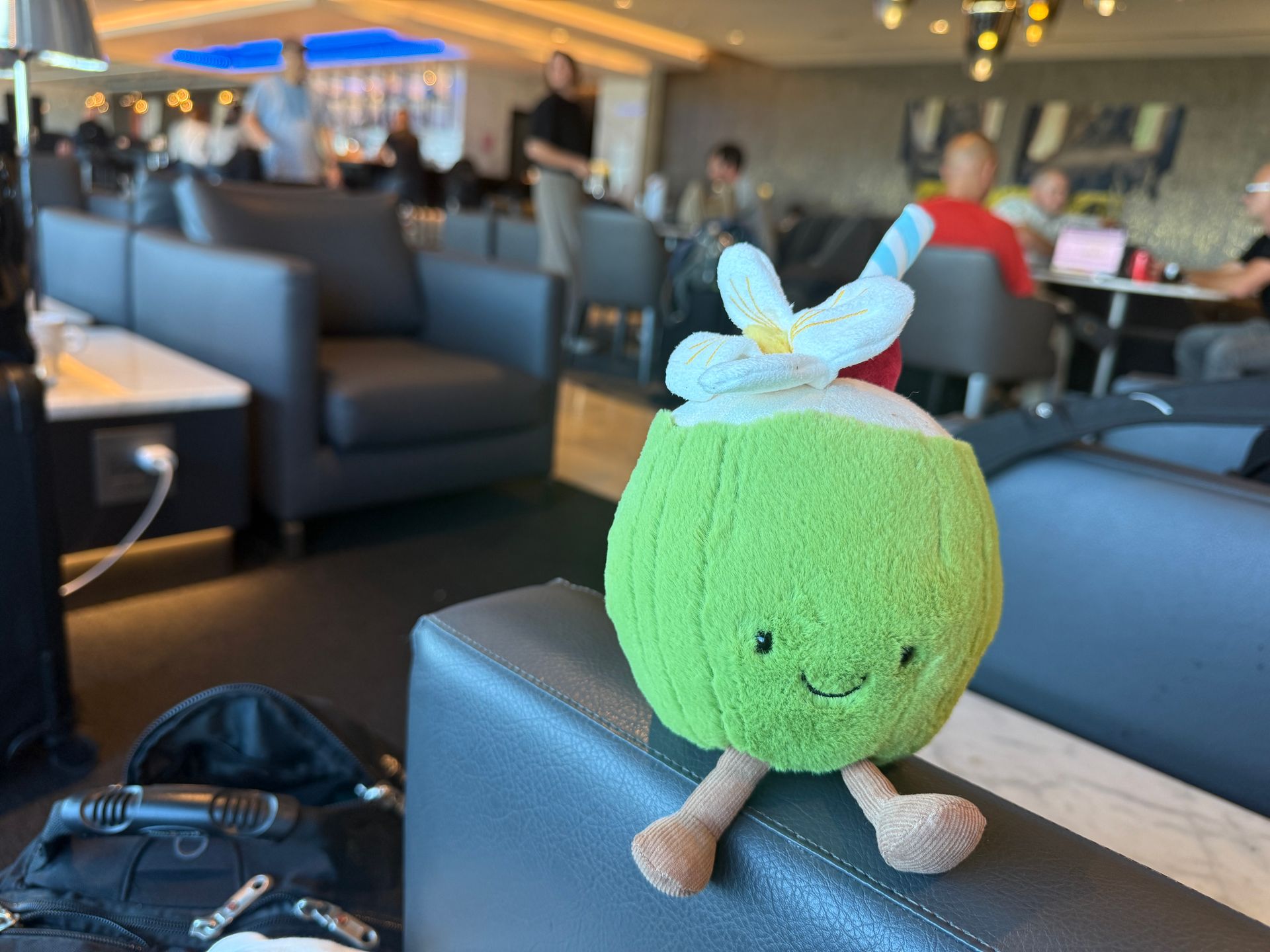 A green coconut plush toy with a straw sits on a black couch in an airport lounge.