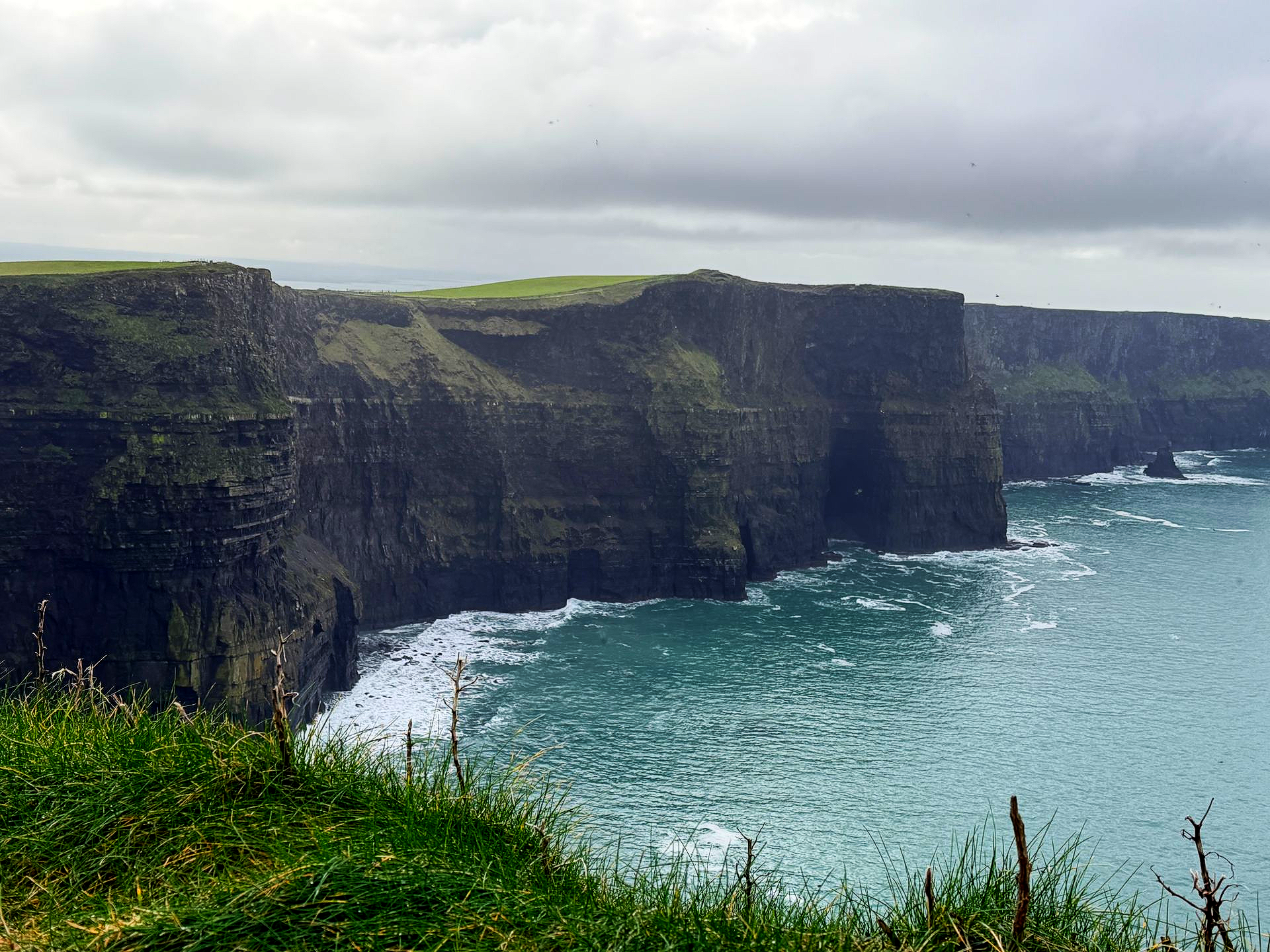 Clouds hang over the green Cliffs of Moher as dark rock walls descend into the blue Atlantic Ocean