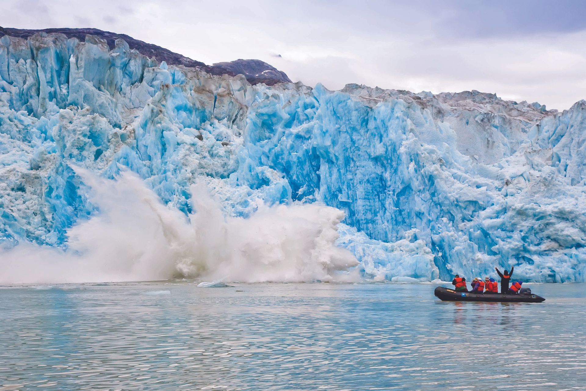 Glacier calving with a large splash in the water. Boat with people watches. Blue ice.