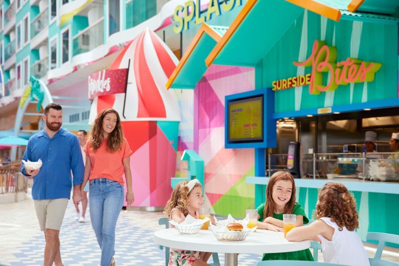 Family walking past a colorful food stand with diners outside; sunny day at a resort.