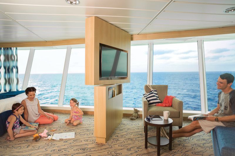 Family relaxing in cruise ship cabin with ocean view; kids play, parent watches TV, other parent reads.