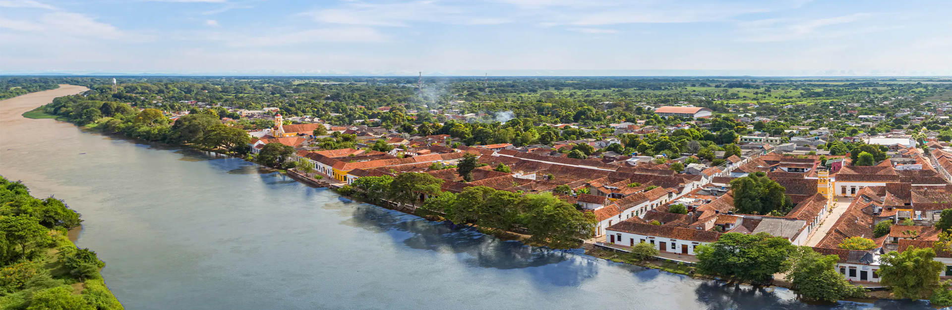 Aerial view of Mompox, Colombia. A river borders a historic town with terracotta rooftops and lush green trees.