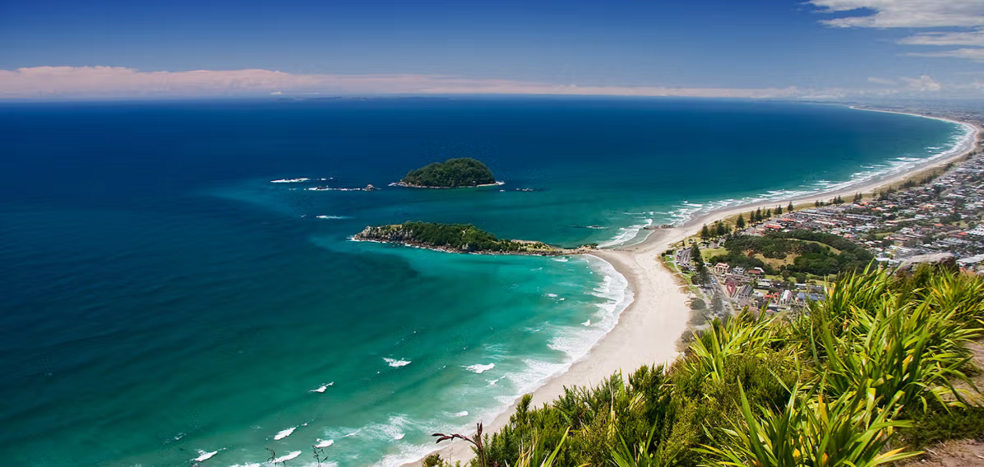 Aerial view of a coastal town with a sandy beach, blue ocean, and a small island under a clear blue sky.