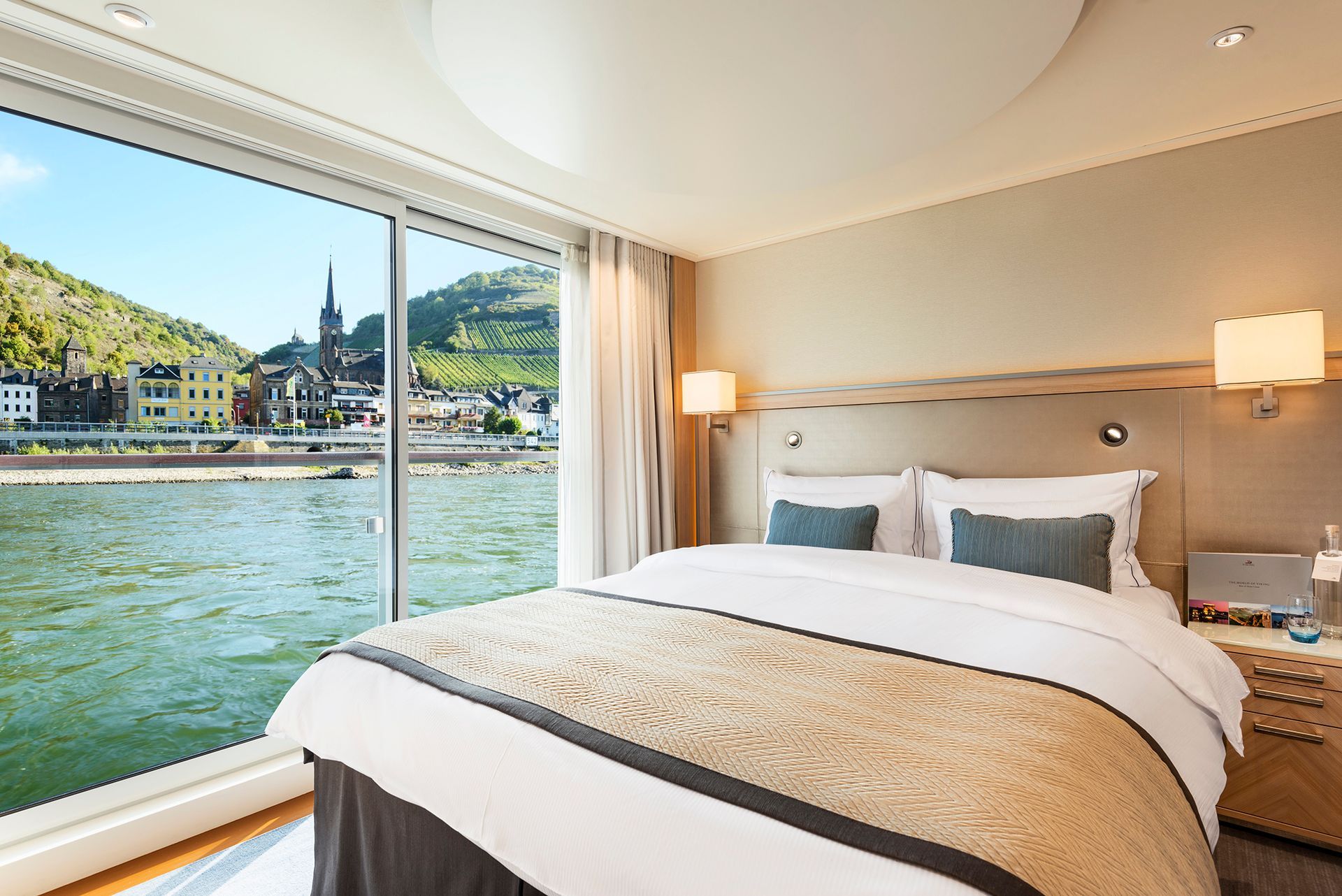 Bedroom with a large window overlooking a river and European town.
