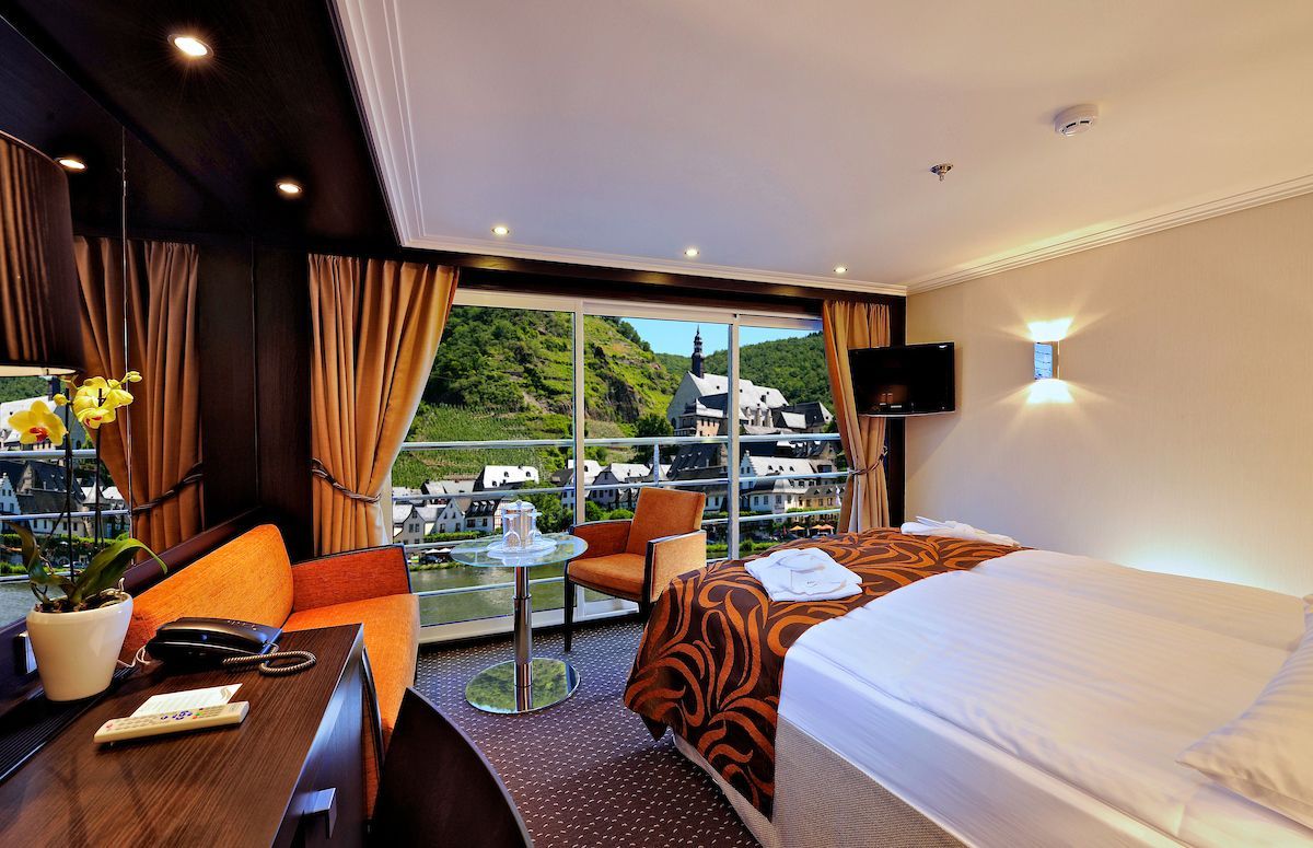Cabin with large window overlooking a town and green hills. Interior with bed, seating, desk, and orange curtains.