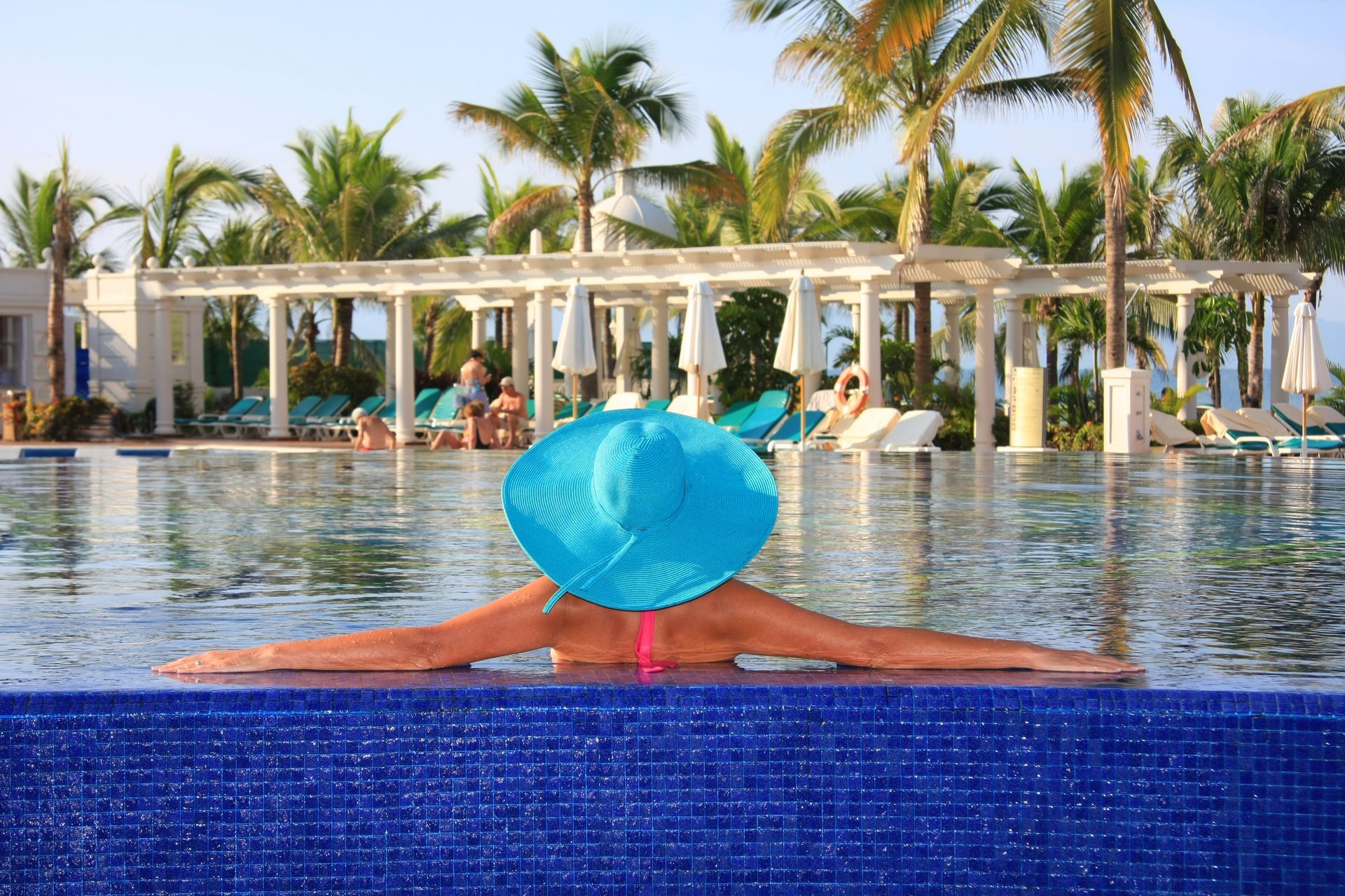 Woman in a blue hat in a pool, arms outstretched. Tropical resort setting with palm trees, blue water, and white structures.