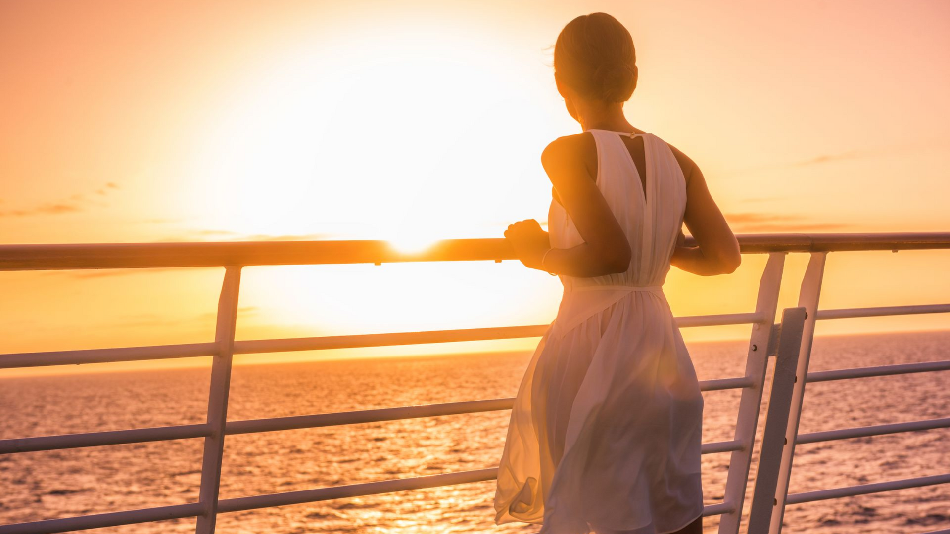 Woman in white dress watches sunset over the ocean from a ship's railing; orange sky.