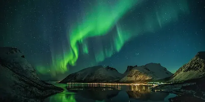 Vibrant green aurora borealis dancing over a snow-covered mountain range and a reflective lake at night.