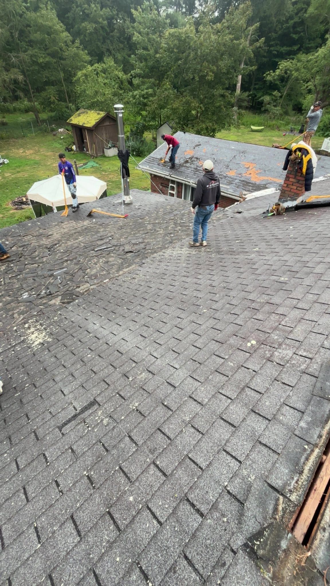 Workers roofing a steep house in the rain, with black shingles, tools, and trees in the background.