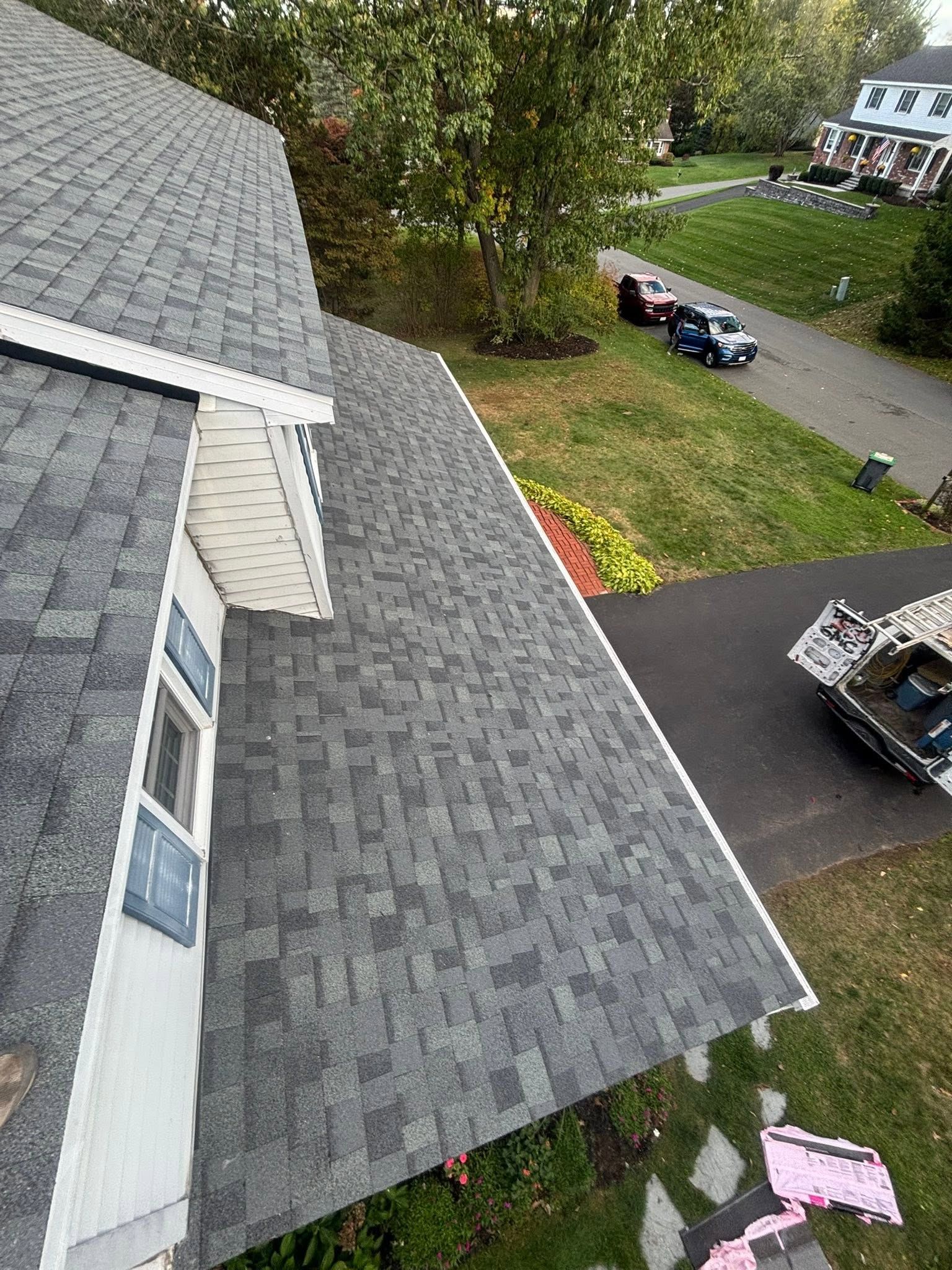 Aerial view of a gray shingled roof beside a driveway with parked vehicles and trees.