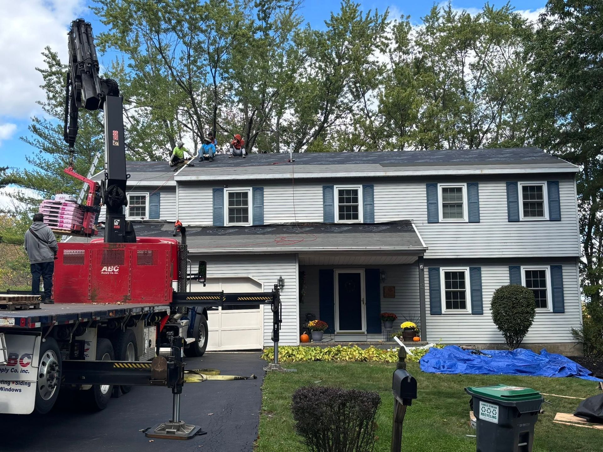 Red utility truck with crane parked by a two-story house; workers on the roof and blue tarp on the lawn