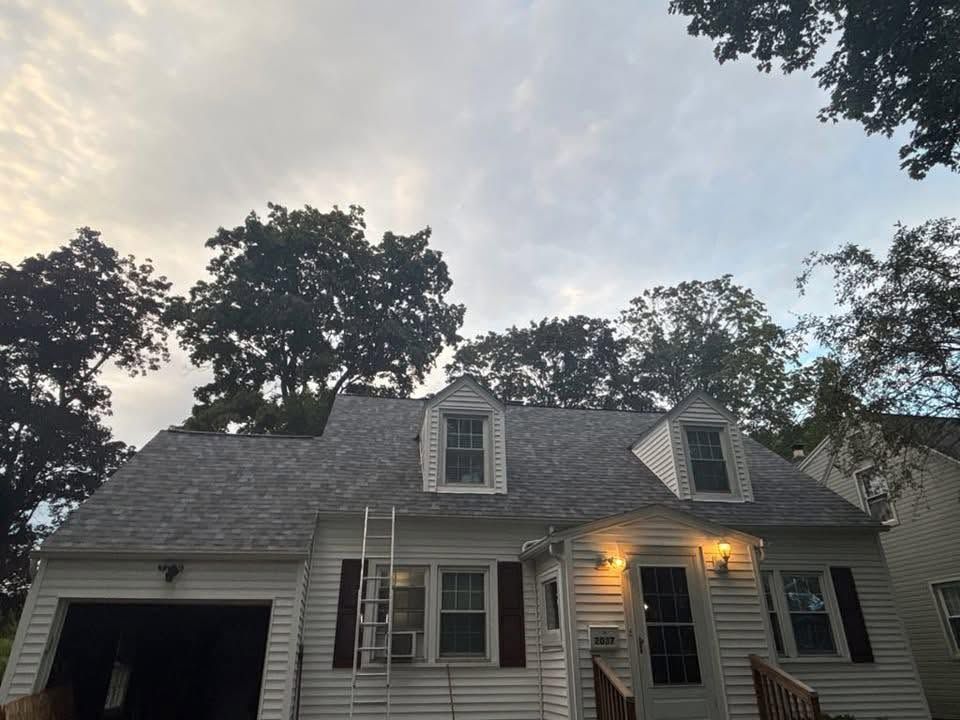 White house with lit porch lights at dusk, framed by tall trees and a cloudy sky