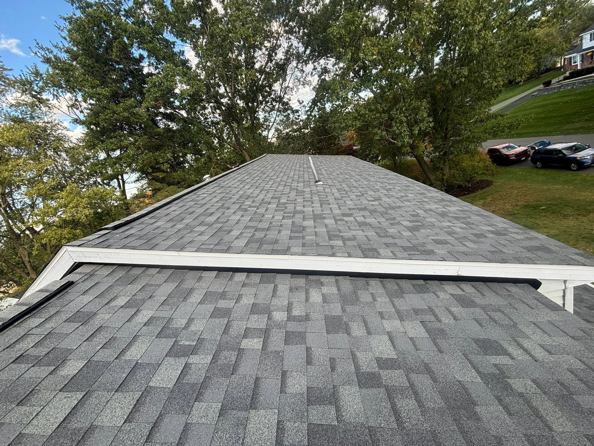 Gray shingled roof with a ridge vent, viewed from above, with trees and parked cars in the background