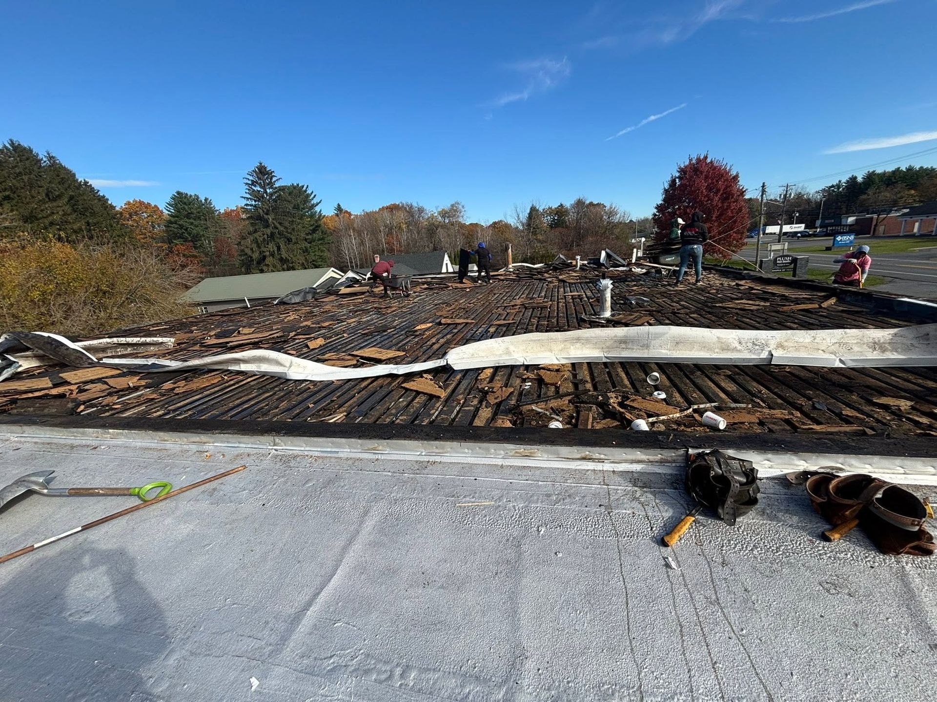 Rooftop in progress with rolled roofing, exposed wood, and workers in the distance under a clear blue sky