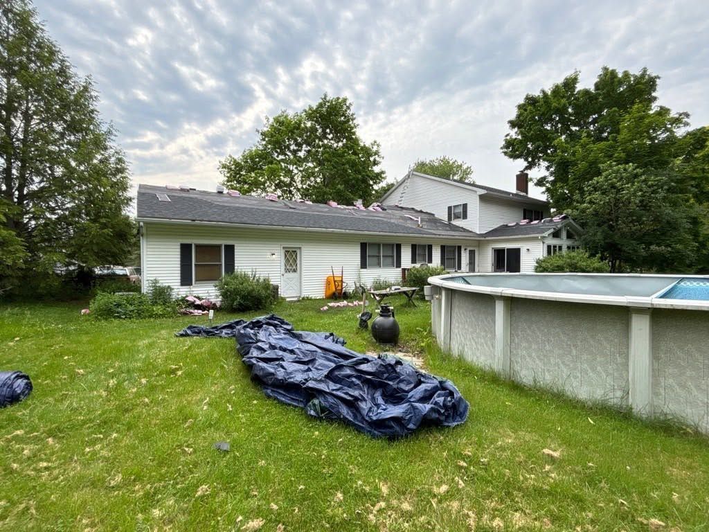 Backyard with a tarp-covered pile, above-ground pool, and a white house under a cloudy sky