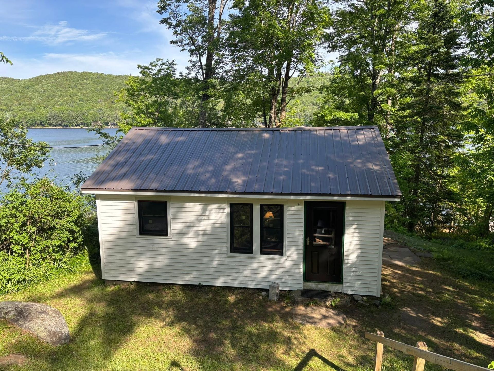 Small white cabin with a dark roof by a lake, surrounded by trees and greenery.