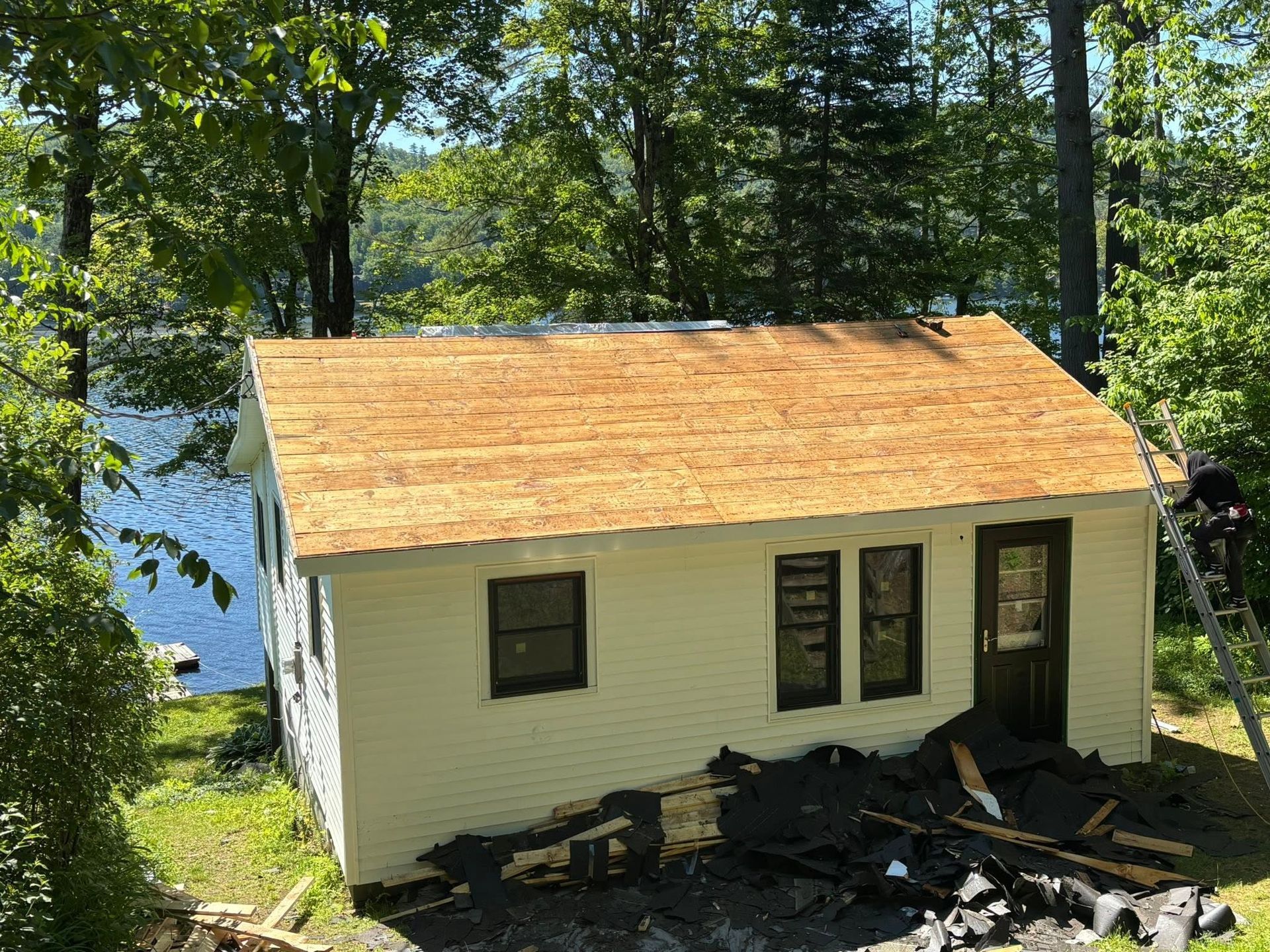 Small white house with a tan roof beside a wooded lake, with debris piled by the front door