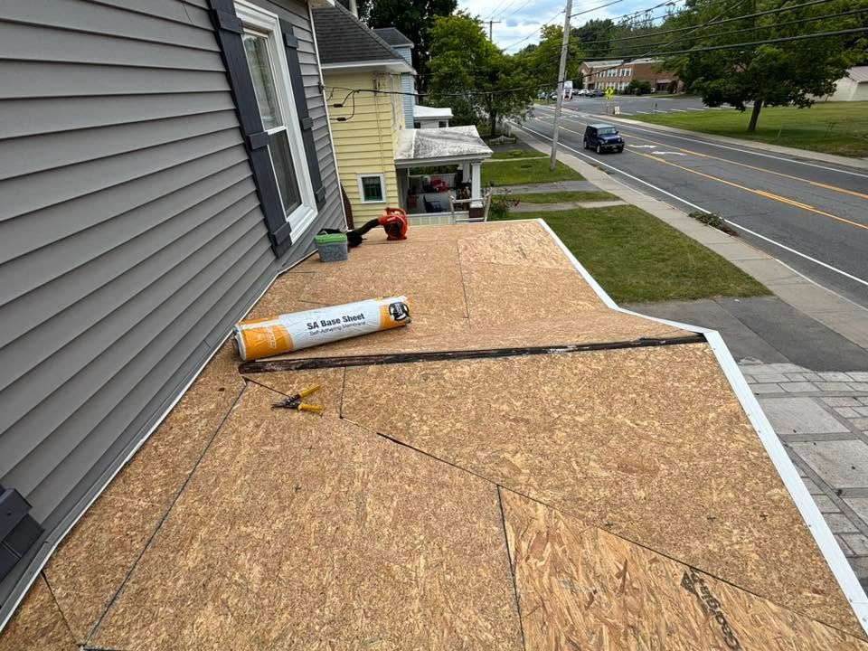 Rooftop under construction with OSB sheathing and a roll of roofing material beside a road