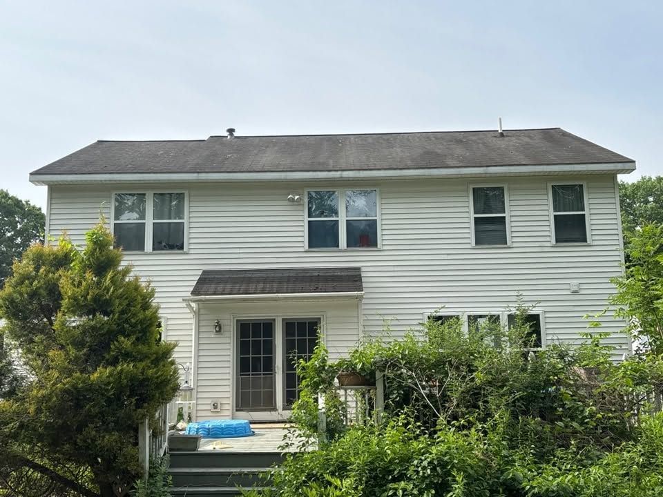 Two-story white house with gray roof, front porch, and dense greenery in the yard