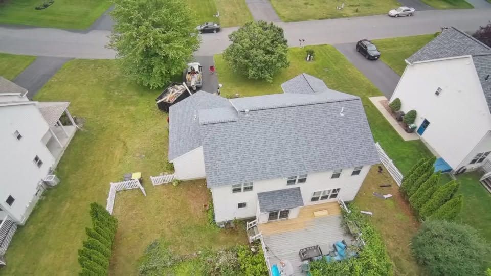 Aerial view of a suburban backyard with a white house, gray roof, driveway, lawn, and neighboring homes.