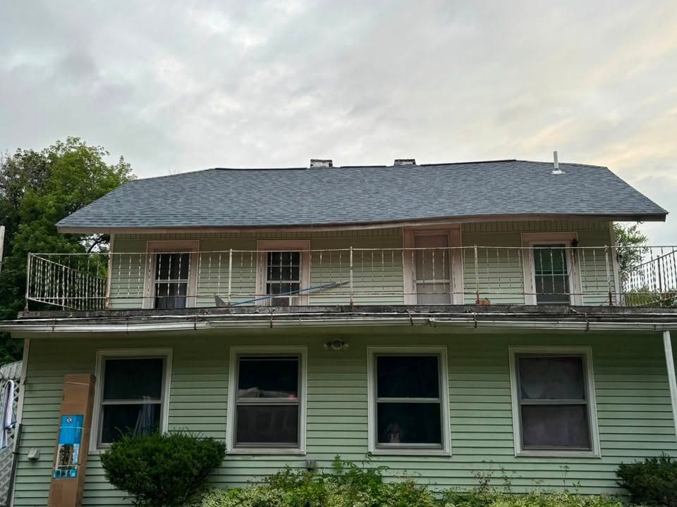 Two-story green house with white trim, front porch, and gray roof under an overcast sky